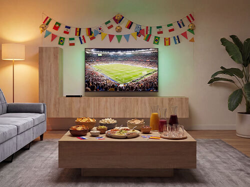 Salon am&eacute;nag&eacute; pour regarder un match de football, avec une t&eacute;l&eacute;vision diffusant une rencontre dans un stade, un canap&eacute; faisant face &agrave; une table basse en bois remplie de collations et de boissons, ainsi que des guirlandes color&eacute;es de drapeaux internationaux d&eacute;corant le mur. Cliquez pour d&eacute;couvrir les produits de la collection.