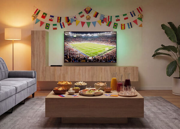 Salon pr&eacute;par&eacute; pour regarder un match de football, avec une t&eacute;l&eacute;vision montrant un stade rempli de spectateurs, un canap&eacute; faisant face &agrave; une table basse en bois garnie de collations et de boissons, ainsi que des drapeaux internationaux color&eacute;s d&eacute;corant le mur.