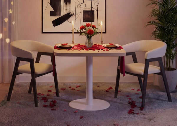 Installation de d&icirc;ner romantique dans un salon moderne avec une petite table ronde blanche et deux chaises rembourr&eacute;es, dress&eacute;e avec des assiettes, des bougies, un vase de roses rouges et des p&eacute;tales de rose &eacute;parpill&eacute;s sur la table et le tapis, encadr&eacute;e par de douces lumi&egrave;res de rideaux, une &oelig;uvre murale abstraite et une plante verte d&rsquo;int&eacute;rieur pour une atmosph&egrave;re cosy de Saint-Valentin.